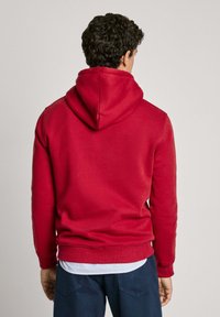 Sudadera roja con capucha de textura suave, con un corte holgado, puños y dobladillo acanalados, superpuesta a una camiseta de rayas.