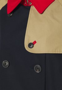 Dubbelknäppt kappa med marinblå kropp, beige krage och röda detaljer. Utmärkande svarta knappar med varumärkeslogotyp och en textur av tyg.