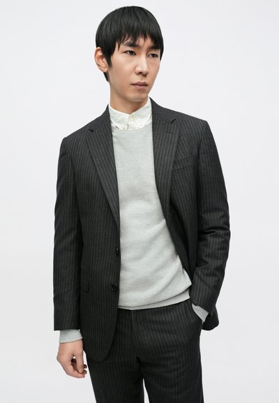 Sivi prugasti odijelo s prilagođenim krojem, slojevito preko svijetlosivog džempera i košulje s ovratnikom, karakterizirano glatkim materijalom i krojem po mjeri.