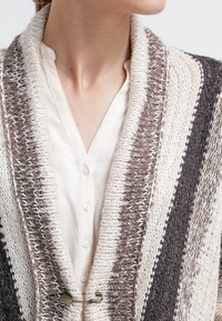 Primer plano de una persona vistiendo una camisa de botones color crema y un cardigan de punto grueso con franjas marrones, beige y crema, sujeto por un gran imperdible.