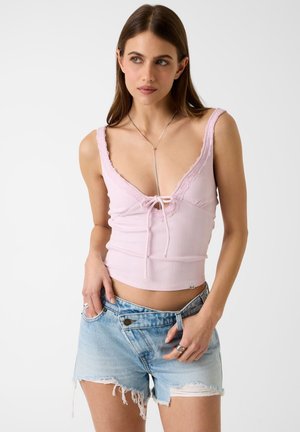 Femme portant un top rose clair sans manches avec bordure en dentelle et détail à nouer, associé à un short en denim clair déchiré et taille haute.