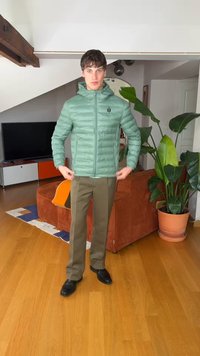 Jeune homme ajustant une doudoune vert clair dans un salon avec un canapé marron, une grande plante en pot, une télévision et un sol en bois.