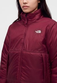 Γυναίκα που φοράει μια μπορντό μπουφάν The North Face με φερμουάρ και ψηλό γιακά, που δείχνει το λογότυπο στο στήθος.