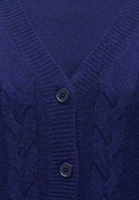 Κοντινός πλάνο ενός ναυτικού μπλε πλεκτού καστόν με ραμμένη σε V λαιμόκοψη, καλωδιακά σχέδια και δύο σκούρα μπλε κουμπιά που είναι κουμπωμένα μπροστά.