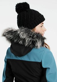 Zwarte gebreide beanie met een pompon, gedragen bij een jas met een blauwe bovenkant en een zwarte onderkant, verfraaid met een rand van nepbont bij de capuchon.