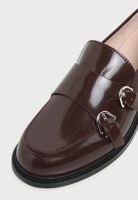 Mocassin en cuir brun foncé brillant avec double boucle argentée et semelle noire sur un fond clair uni.