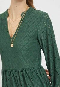 Grön klänning i strukturerad tyg med en rynkad midja, långa ärmar och utskurna mönster. Den är kombinerad med lager av guldkedjor.