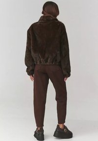 Brązowa kurtka z futra ekologicznego z wysokim kołnierzem, marszczoną talią i długimi rękawami; zestawiona z brązowymi dopasowanymi spodniami i czarnymi butami na platformie.