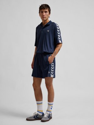 En ung mand står iført navy blå atletisk polo og shorts med hvide chevronstriber, hvide sokker med blå og gule striber og grå sneakers.