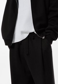 Schwarze Zip-Jacke mit geripptem Kragen über einem weißen strukturierten Pullover, kombiniert mit schwarzen Jogginghosen mit Kordelzug und seitlichen Taschen.