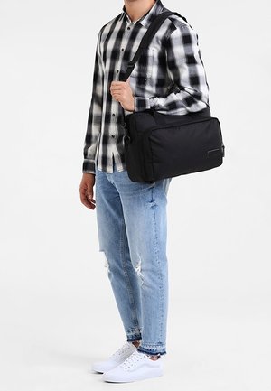 Man draagt een zwart-wit geruit overhemd en gescheurde blauwe spijkerbroek, draagt een zwarte schoudertas en witte sneakers.