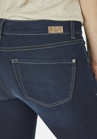 Tmavě modré denimové džíny s zadní kapsou a hnědou koženou etiketou s nápisem "Angels" a viditelnými švy.