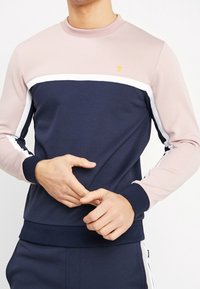 Sudadera de cuello redondo con un cuerpo azul marino, hombros rosas y franjas blancas. Presenta un pequeño logo y puños y dobladillo acanalados.