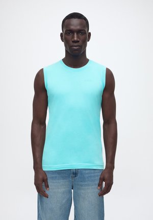 Muskulárny muž v svetlomodrom tielku bez rukávov a svetlomodrých džínsoch stojí pred jednoliatym bielym pozadím.