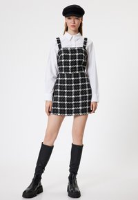 Vestido mini a cuadros en blanco y negro con tirantes finos, tejido texturizado, combinado con una camisa blanca y botas negras hasta la rodilla.