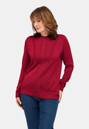 Maglione rosso lavorato a maglia con collo a giro, caratterizzato da un design a trecce testurizzate lungo il centro e bordo e polsini a coste. Abbinato a jeans blu.