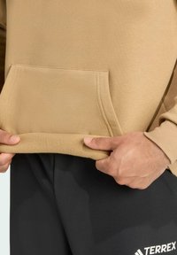 Bež dukserica s prednjim džepom, koja ima meku teksturu i rub obložen flisom. Uparena s crnim hlačama koje prikazuju sportski dizajn.