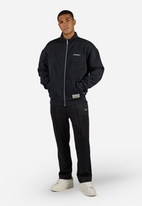 Sort fleece zip-up jakke med høj krave, prydet med et logo på brystet; kombineret med sorte bukser og lysfarvede sneakers.