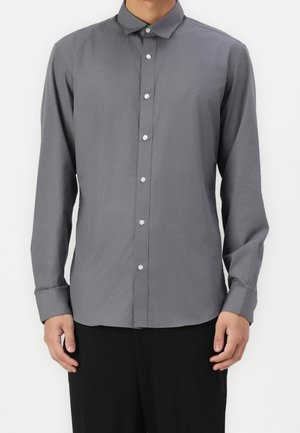Homme portant une chemise grise unie à manches longues boutonnée et un pantalon noir, debout devant un fond blanc.