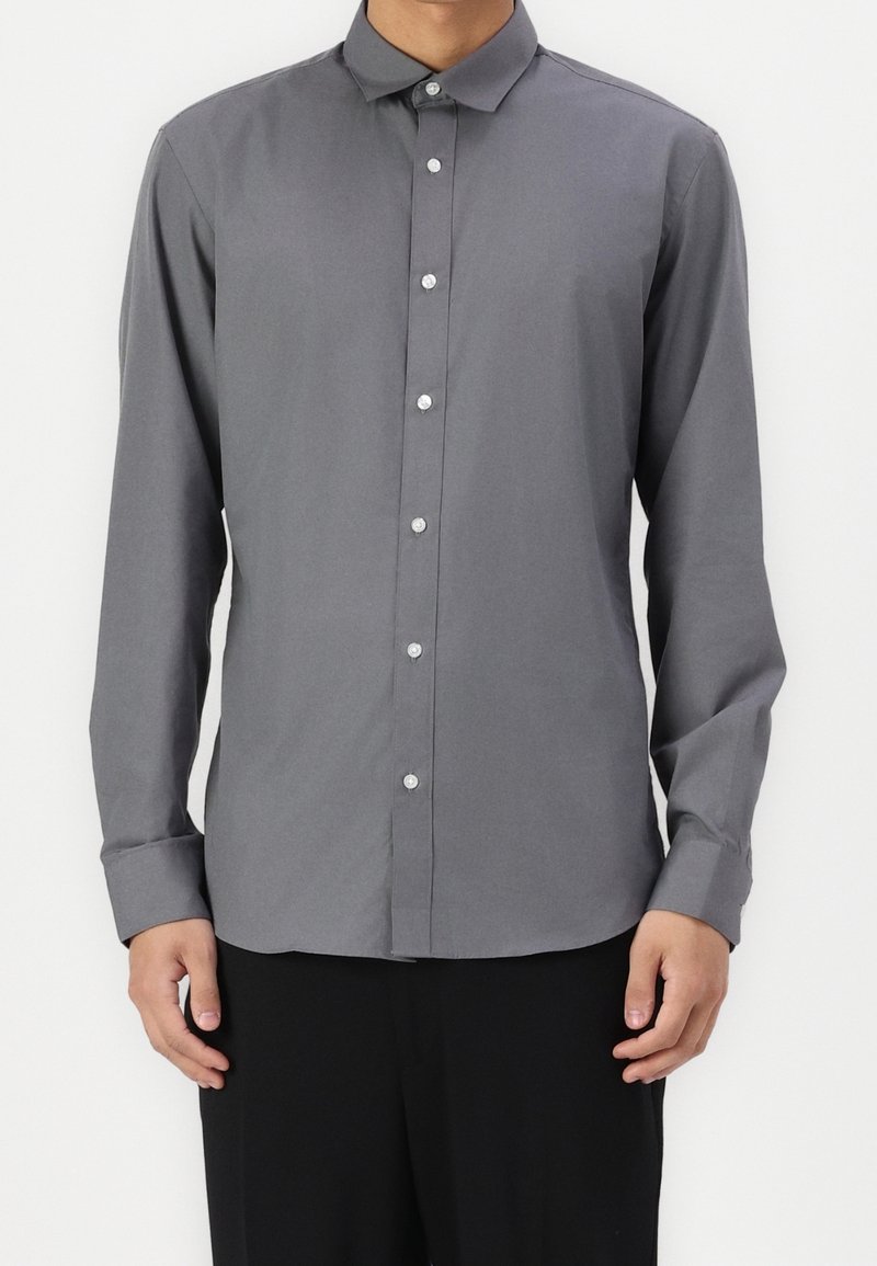 Homme portant une chemise grise unie à manches longues boutonnée et un pantalon noir, debout devant un fond blanc.
