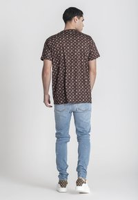 Lichtbruine shirt met korte mouwen met een herhalend logo patroon, gecombineerd met lichtblauwe distressed jeans en bruin met patroon sneakers.