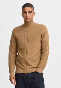 Tamni zip-up pulover s visokim ovratnikom, teksturiranim pletenim uzorkom, rebrastim manžetama i rubom. Nošen preko tamnih hlača s prilagođenim krojem.