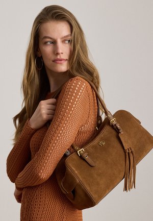 Femme aux longs cheveux portant un haut en maille couleur rouille, tient un sac à main en daim marron avec ferrures dorées et pompons sur son épaule.