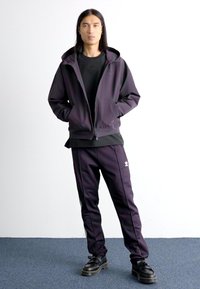Dark purple athletic tracksuit with zip-up hoodie, black t-shirt, and stripes. Features logo on pants. Smooth texture, casual fit.