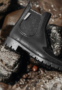 Zwarte rubberen Chelsea-boot met een getextureerd elastisch paneel, voorzien van een robuuste zool en waterdruppels, gepositioneerd op natte stenen.