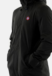 Veste noire imperméable avec capuche, arborant un logo circulaire rouge et bleu sur la poitrine ainsi que des poches latérales. Texture lisse, design moderne.