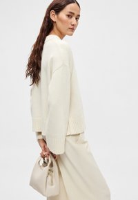Krém színű kötött pulóver bordázott mandzsettával, világos bézs színű, széles szárú nadrággal párosítva. A modell egy kis bézs táskát tart, arany cipzáras részlettel.