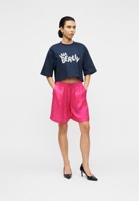 Ναυτικό μπλε κοντομάνικο μπλουζάκι με λευκό κείμενο και ροζ σατέν σορτς με κορδόνι στη μέση, συνδυασμένα με μαύρες ψηλοτάκουνες γόβες με μυτερή μύτη.