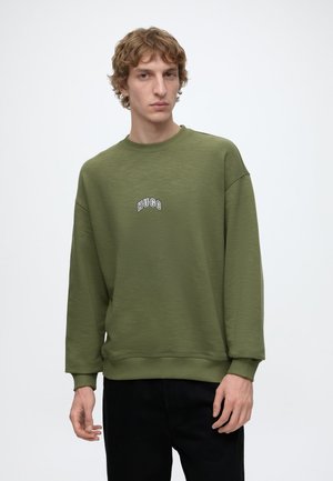 Young man with curly hair wearing an olive green sweatshirt with "HUGO" text on the chest and black pants against a plain white background.