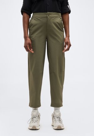 Persoon draagt olijfgroene taps toelopende broek, beige sneakers en een zwart shirt met lange mouwen, staand tegen een effen lichte achtergrond.