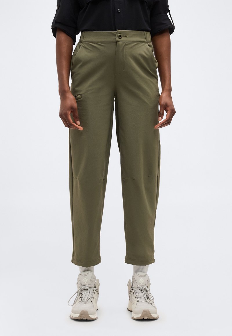 Άτομο που φοράει παντελόνια ελιάς στενά στο κάτω μέρος, μπεζ αθλητικά παπούτσια και μαύρο μπλουζάκι με μακριά μανίκια, στέκεται μπροστά σε ένα απλό φωτεινό φόντο.