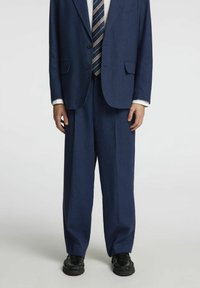 Costume bleu marine avec un blazer à deux boutons et un pantalon à jambes larges, en tissu texturé, associé à une cravate rayée et des chaussures noires brillantes.
