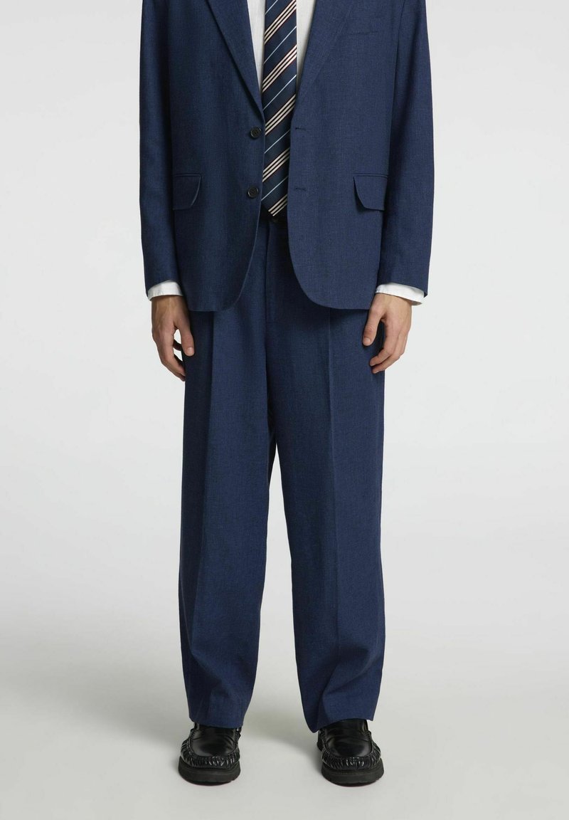 Costume bleu marine avec un blazer à deux boutons et un pantalon à jambes larges, en tissu texturé, associé à une cravate rayée et des chaussures noires brillantes.