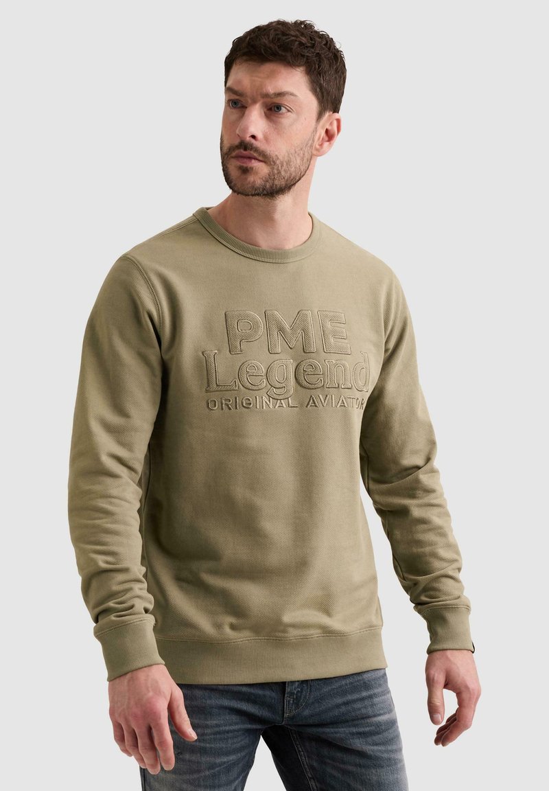 Άνδρας που φοράει ελιά πράσινο φούτερ PME Legend και μπλε τζιν, στέκεται μπροστά σε απλό ανοιχτό φόντο, κοιτάζοντας προς τα πλάγια.