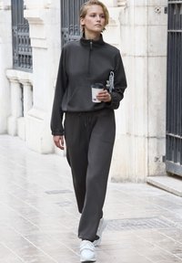 Felpa zip grigia scura con colletto alto e tasca frontale, abbinata a pantaloni sudati con gamba tapered e sneakers bianche.