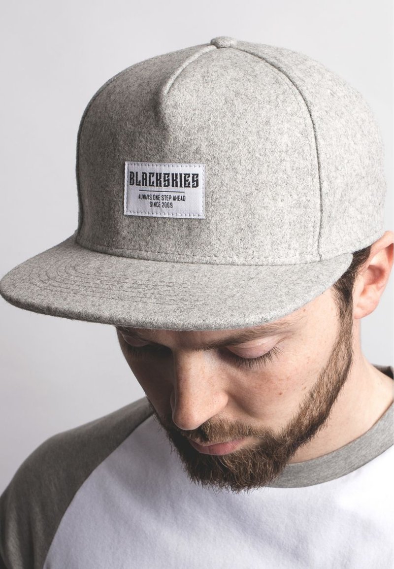 Siva kapa s sivo plosko obrobo in z nakitom logotipom "BLACKSKIES" na sprednji strani. Mehka tekstura in zaobljena oblika krošnje.