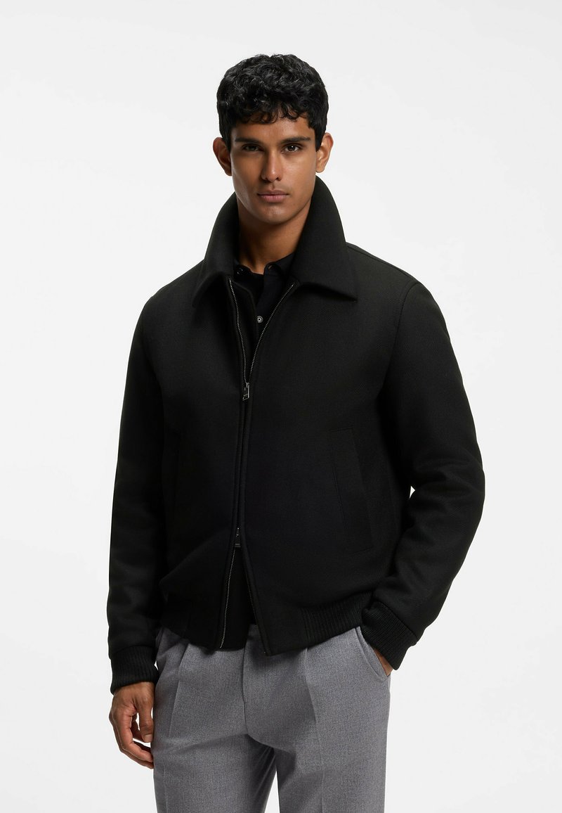 Young man with short curly hair wearing a black zip-up jacket with a wide collar and gray pants, standing with one hand in pocket.