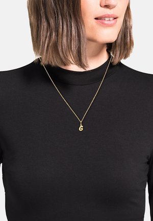 Goldkette mit einem Anhänger in Form des Buchstabens "G", getragen zu einem schwarzen Hochkragenoberteil. Die Kette ist fein und zart und ergänzt das schlichte Design.