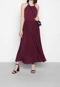 Burgundyröd plisserad maxikjol med halterneckdesign, gjord av mjukt tyg; ihop med svarta klacksandaler och en liten svart väska.