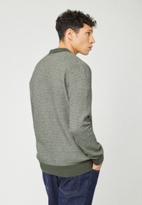 Man met kort krullend haar, in een groen geruit sweater en blauwe jeans, staat met zijn handen in zijn zakken, zijwaarts gericht tegen een effen achtergrond.