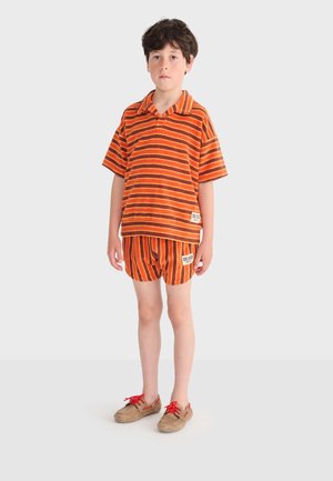 Garçon debout portant un t-shirt à manches courtes rayé orange et marron et un short assorti avec des chaussures beige à lacets rouges, fond neutre.