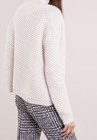 Pull blanc en tricot avec un motif texturé, col montant et poignets côtelés, assorti à un pantalon à carreaux noirs et blancs.