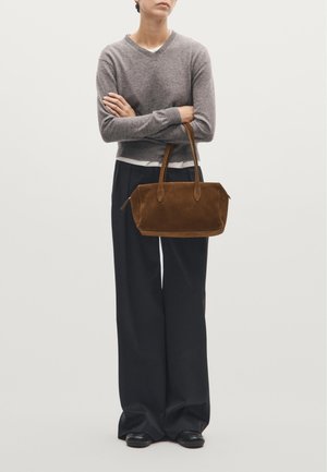 Hnědá semišová kabelka s kulatými hranami, dvojitými uchy a hladkým povrchem. Nosená s šedým svetr s véčkovým výstřihem a širokými černými kalhotami.