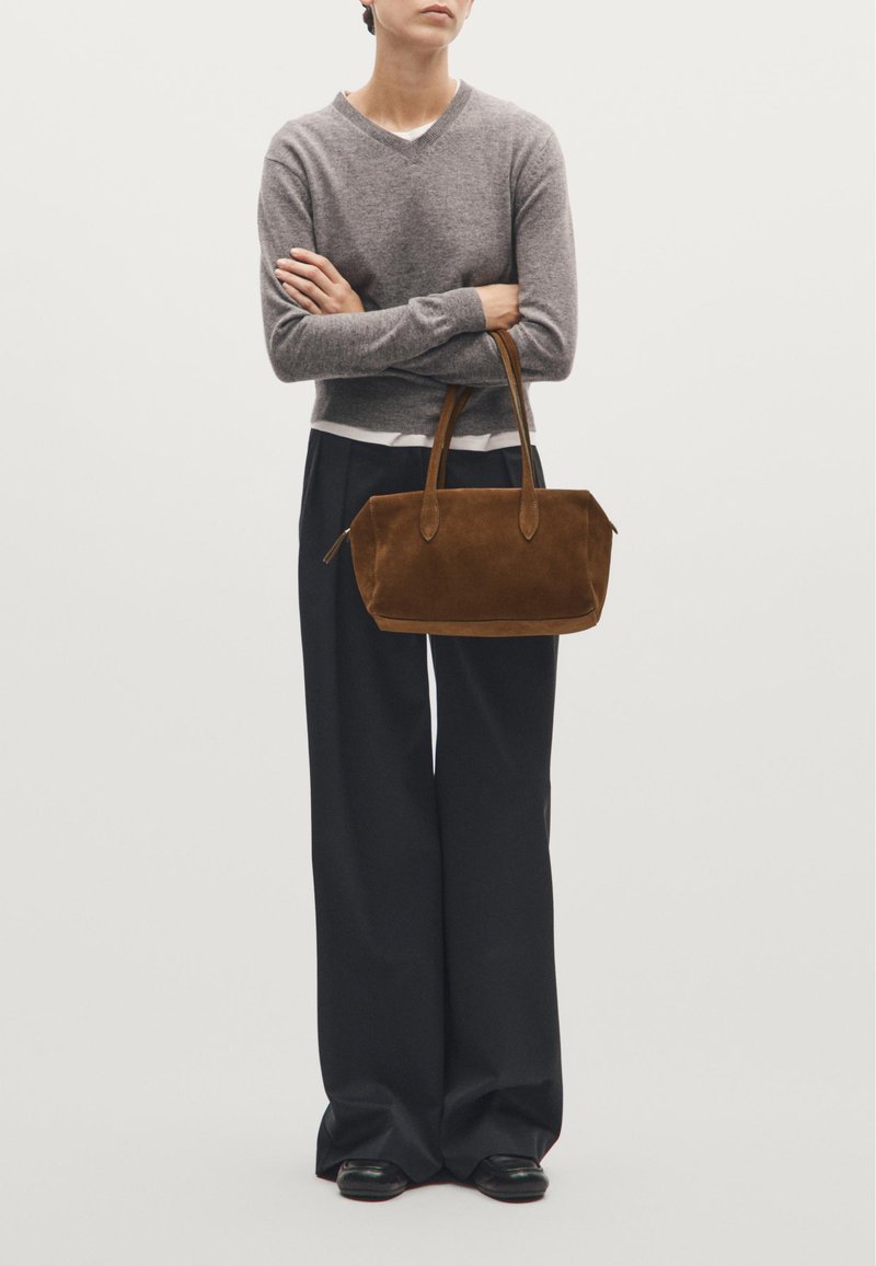 Suedes håndtaske i brun med afrundede kanter, dobbelte håndtag og en glat tekstur. Bæres med en grå V-hals sweater og brede sorte bukser.