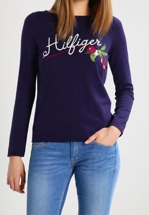 Vrouw die een marineblauw langärmelig shirt draagt met de tekst "Hilfiger" en bloemrijke borduurwerk, gecombineerd met lichtblauwe spijkerbroek, staand met één hand in de zak.