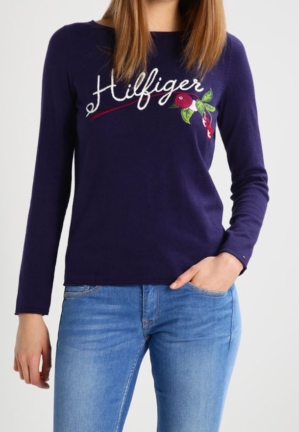 Femme portant un haut à manches longues bleu marine avec le texte « Hilfiger » et une broderie florale, associée à un jean bleu clair, debout avec une main dans la poche.